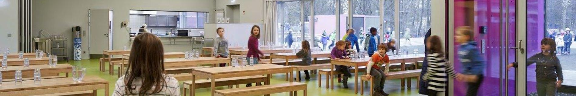 Schulmensa mit Tischen und Bänken, im Hintergrund die Essensausgabe. Schülerinnen und Schüler kommen zur Tür herein.